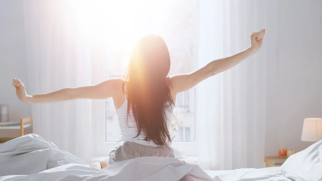 Woman stretching while waking up in bed with morning sunlight